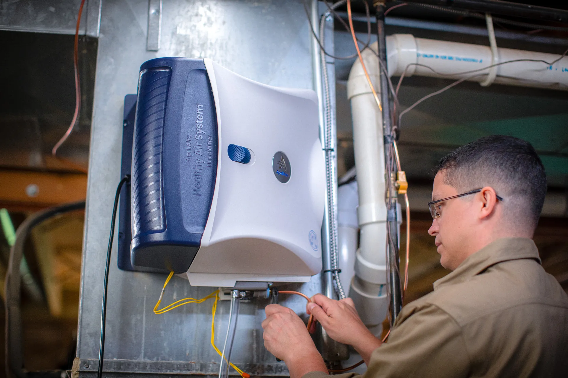 A technician adjusts a white AprilAire whole-home air quality unit inside an HVAC cabinet, with ducts and wiring nearby.