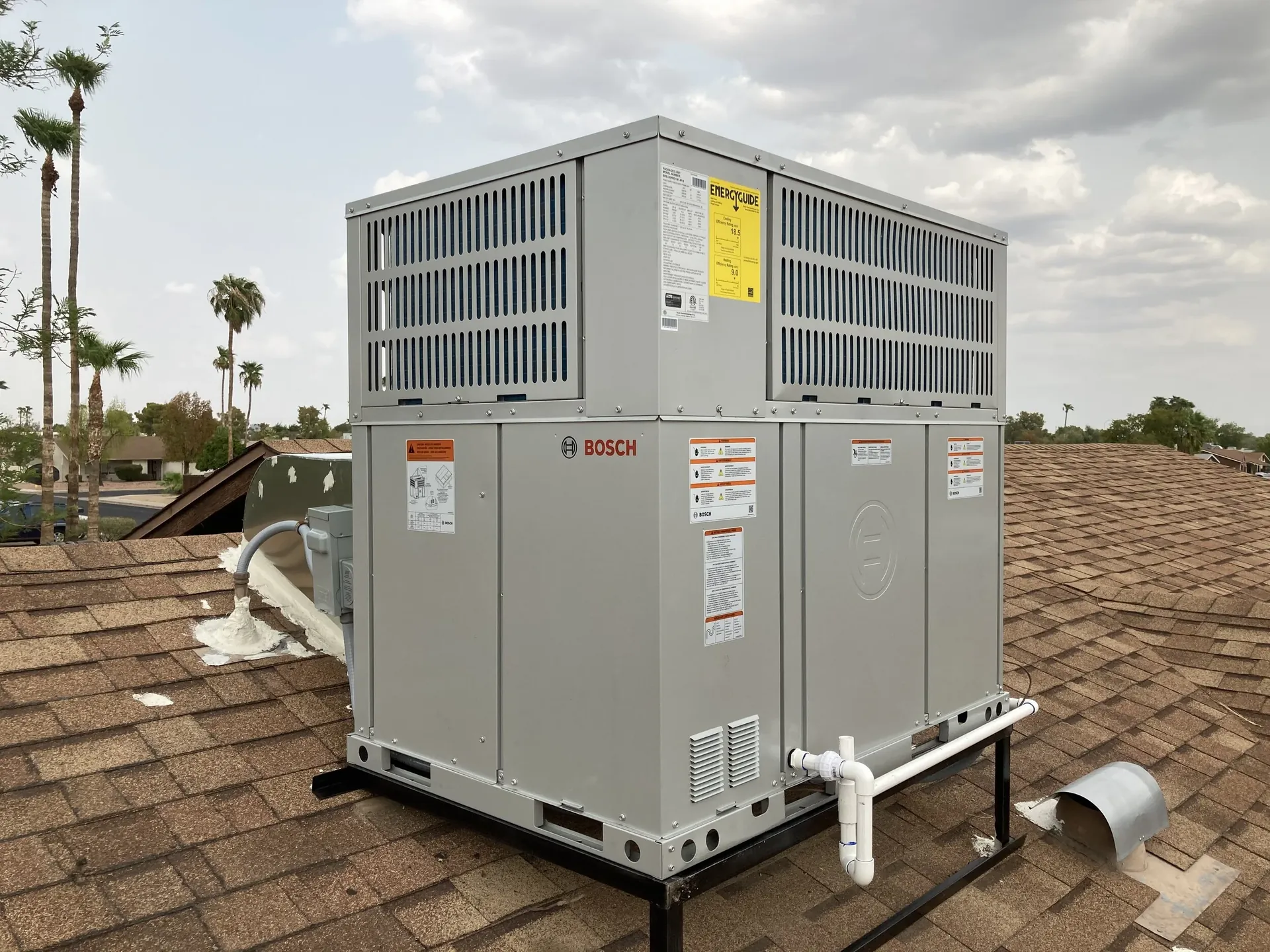 A gray Bosch HVAC rooftop unit with vented metal panels and white drain piping stands on a shingled roof under a cloudy sky.