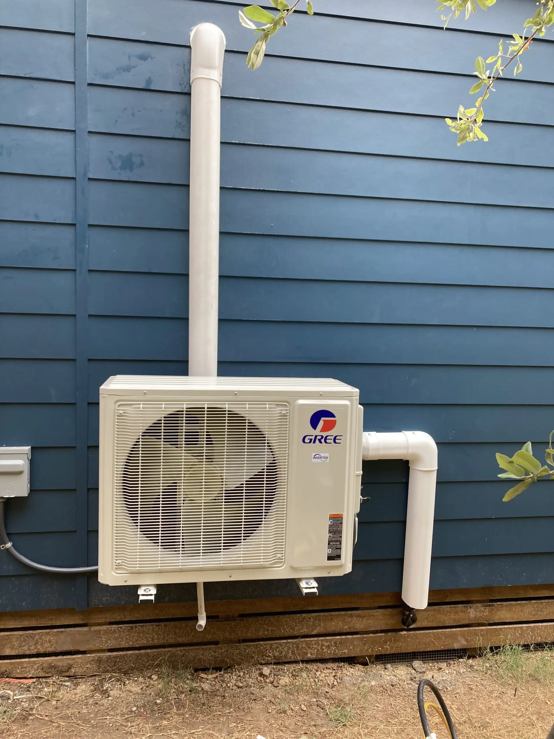 A cream-colored GREE outdoor HVAC unit with a large front fan stands against dark blue siding, connected to white piping.
