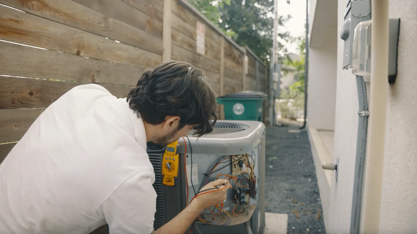 A technician in a white shirt tests the exposed wiring and controls of an outdoor HVAC unit beside a wooden fence in Austin.