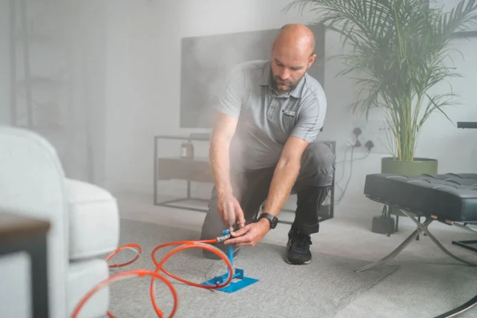 A technician kneels on a gray carpet and connects an orange hose to a blue floor-level device inside a bright home.