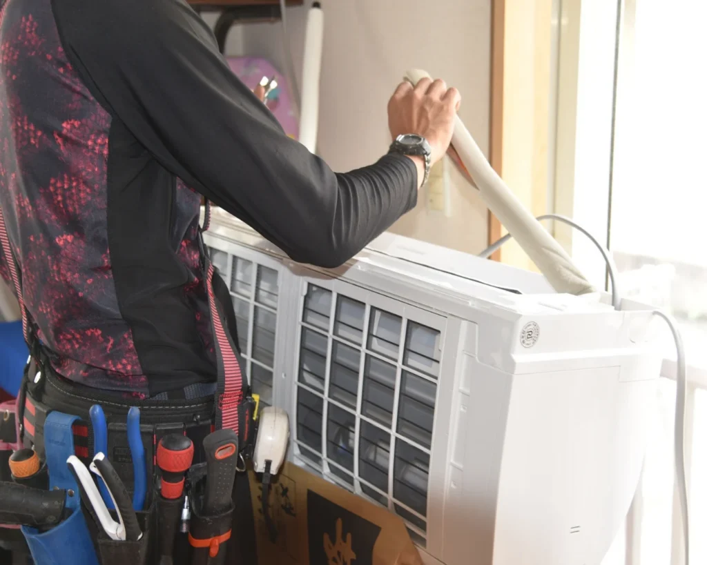 A technician in dark workwear reaches up to adjust a white HVAC unit, with a tool belt and equipment panels visible nearby.