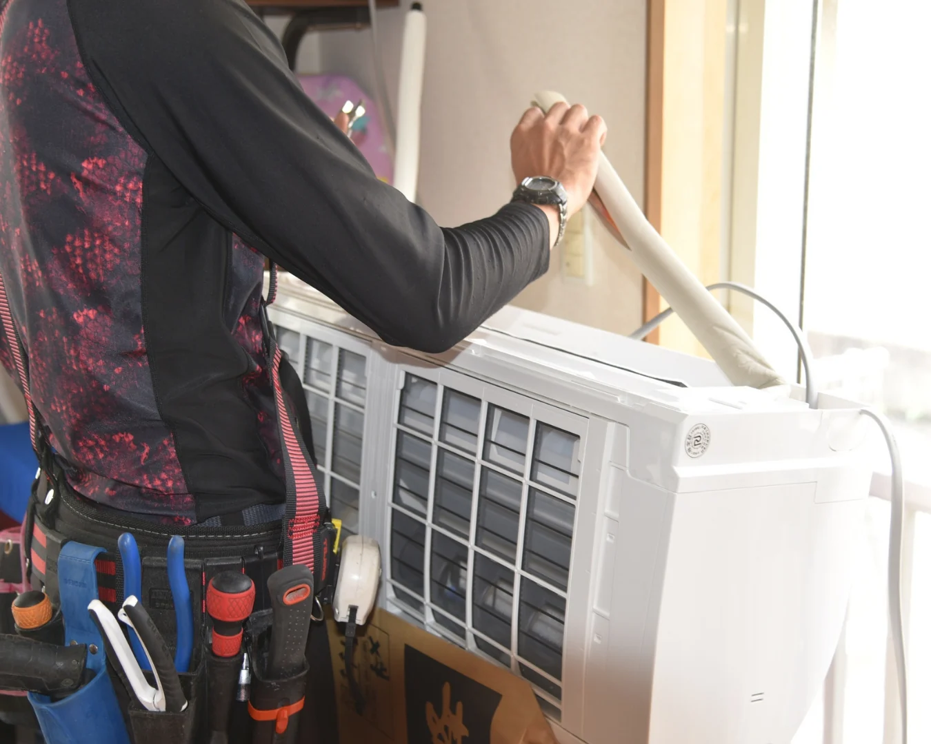 A technician in dark workwear reaches up to adjust a white HVAC unit, with a tool belt and equipment panels visible nearby.