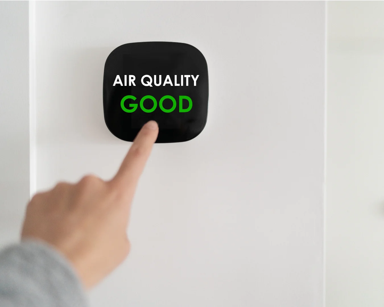 A fingertip presses the green “GOOD” indicator on a black air quality monitor mounted against a plain white wall.