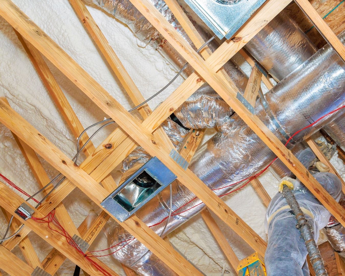 Insulated HVAC ductwork runs through a timber-framed attic, with silver flexible ducts, wiring, and a ventilation unit visible.