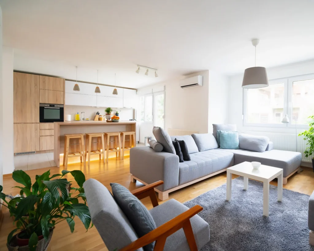 A bright open-plan living room with a gray sectional sofa, wooden kitchen cabinetry, a breakfast bar, and large windows.