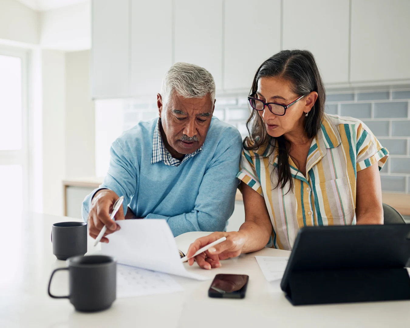 An older man and a woman in glasses review paperwork together at a desk, with a tablet, pens, and coffee cups nearby.
