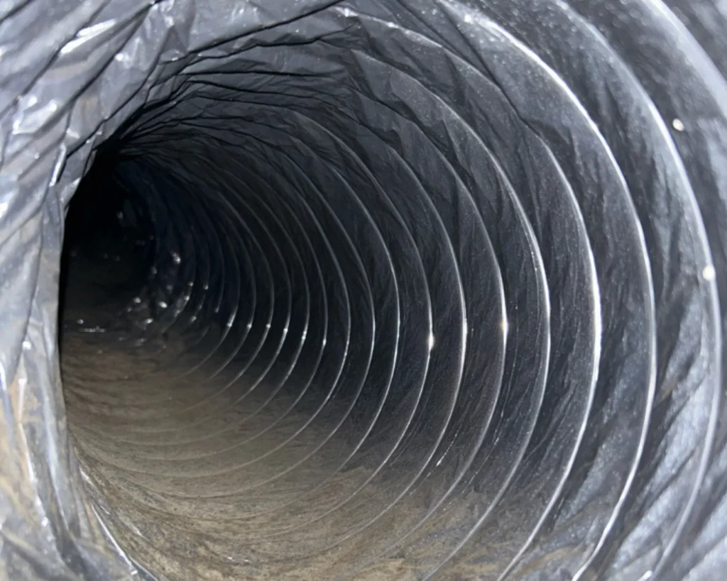 A close view inside a dark, ribbed HVAC duct shows dust and light-colored residue along the lower surface and inner folds.