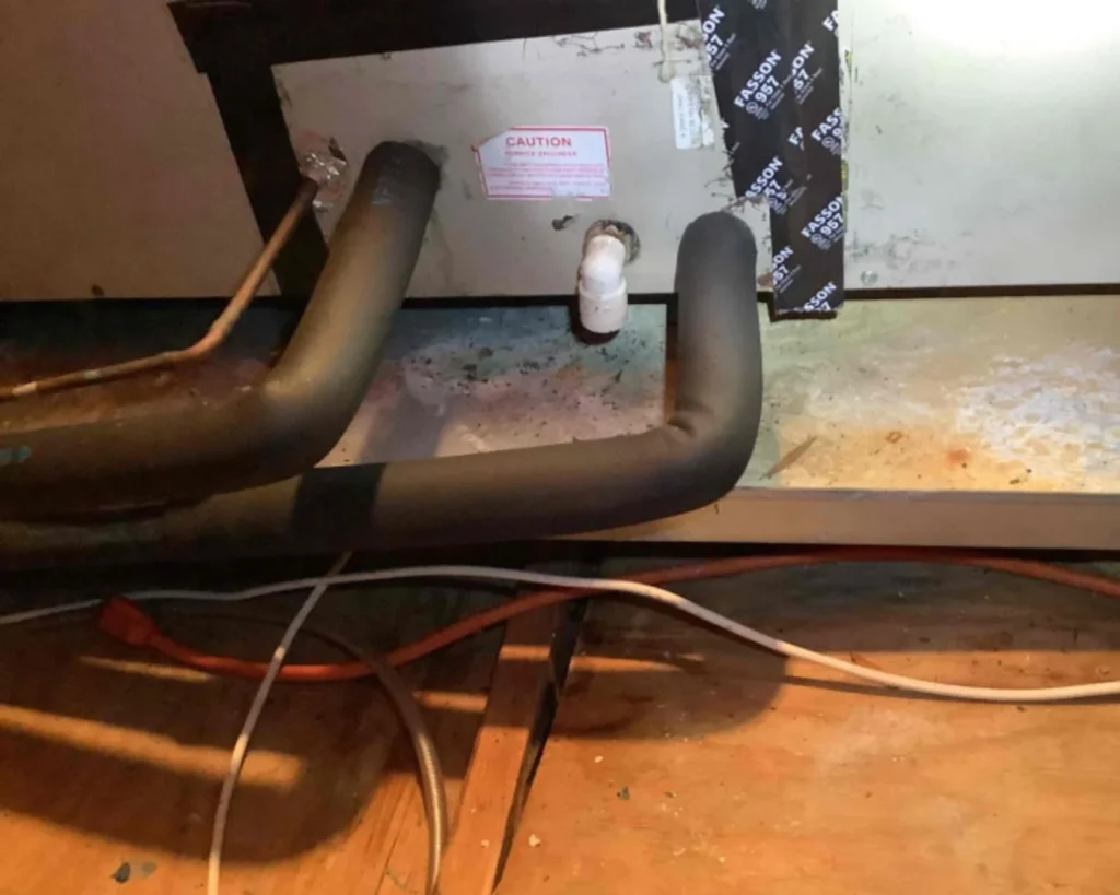Large insulated HVAC pipes and smaller utility lines run through a tight attic space, with a caution label visible on the panel above.