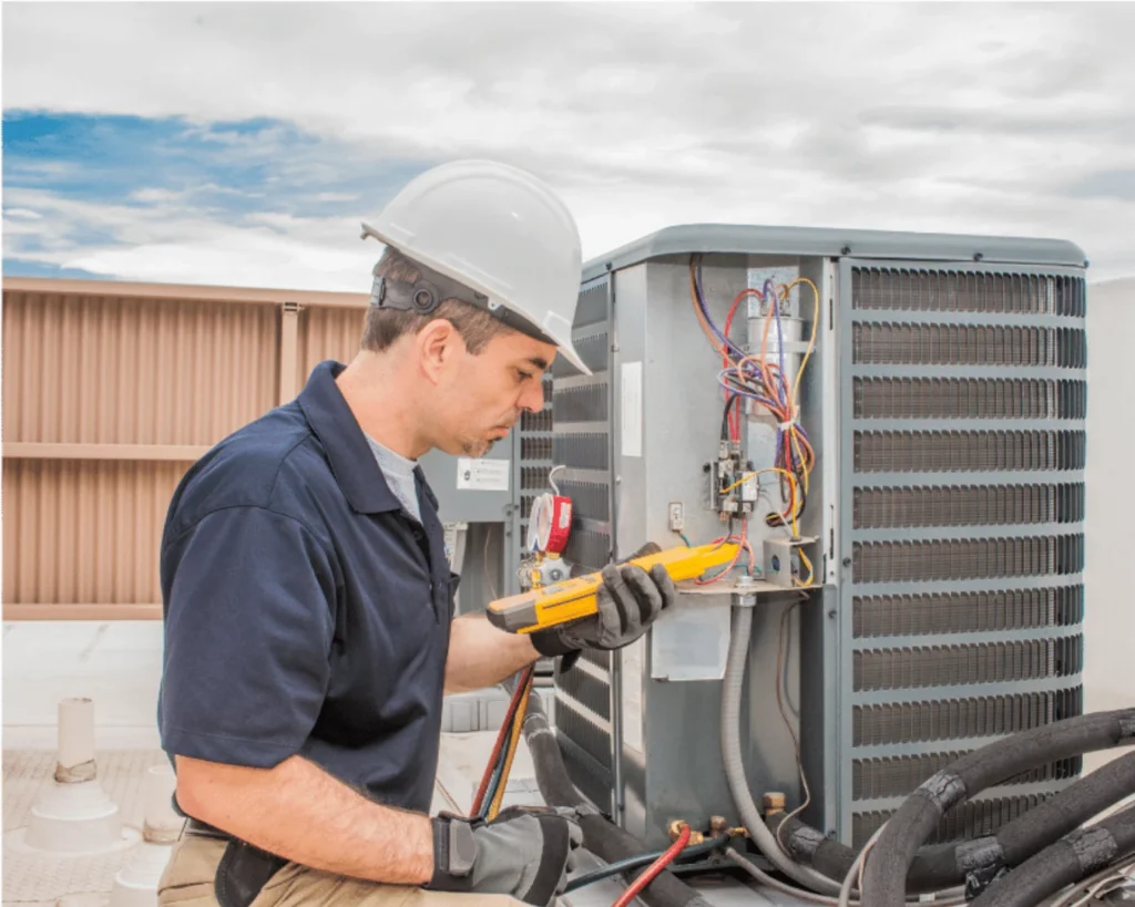 A technician in a white cap and dark uniform inspects exposed wiring inside a gray outdoor HVAC unit with a yellow tool.