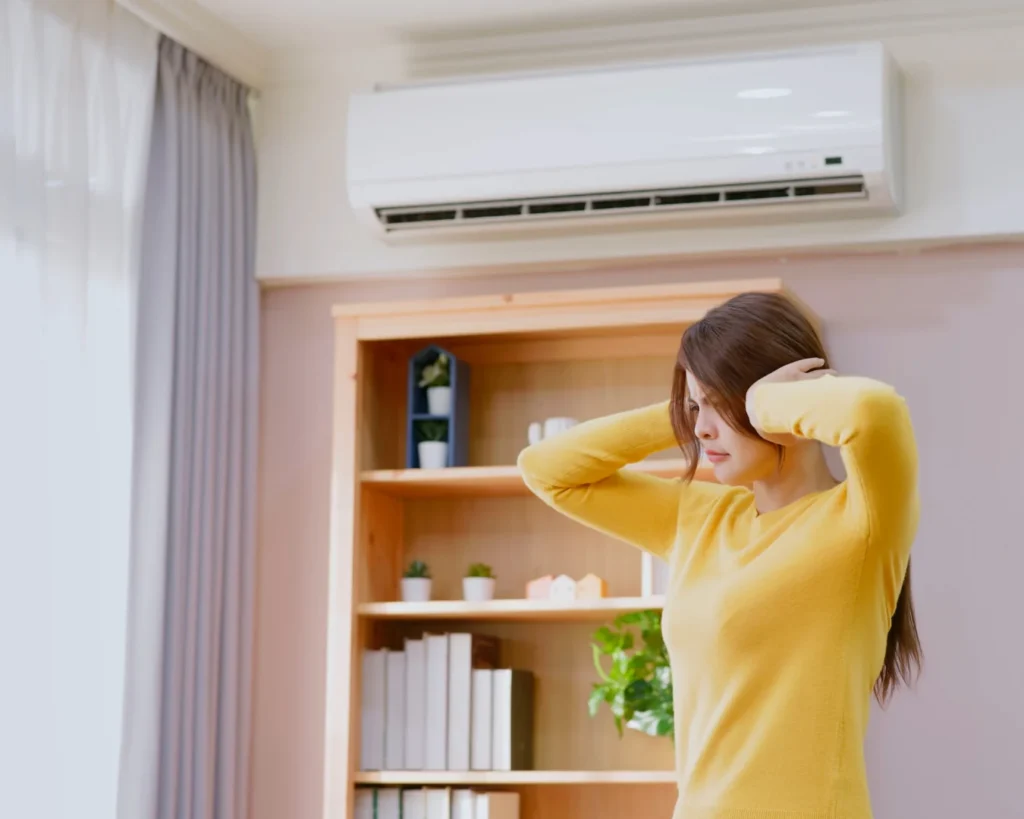 A woman in a yellow sweater covers her ears beneath a wall-mounted air conditioner, reacting to a loud sound in a home.