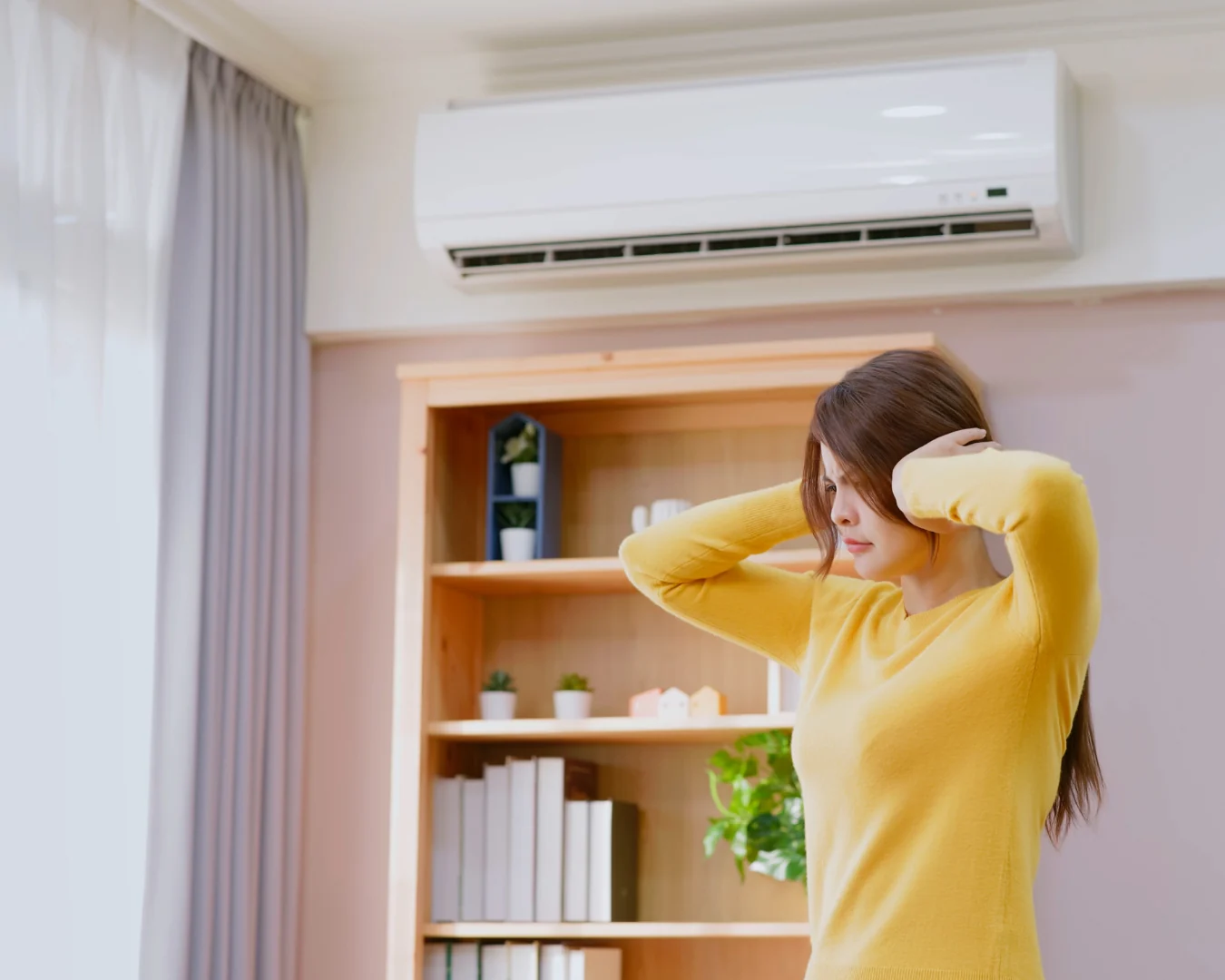 A woman in a yellow sweater covers her ears beneath a wall-mounted air conditioner, reacting to a loud sound in a home.