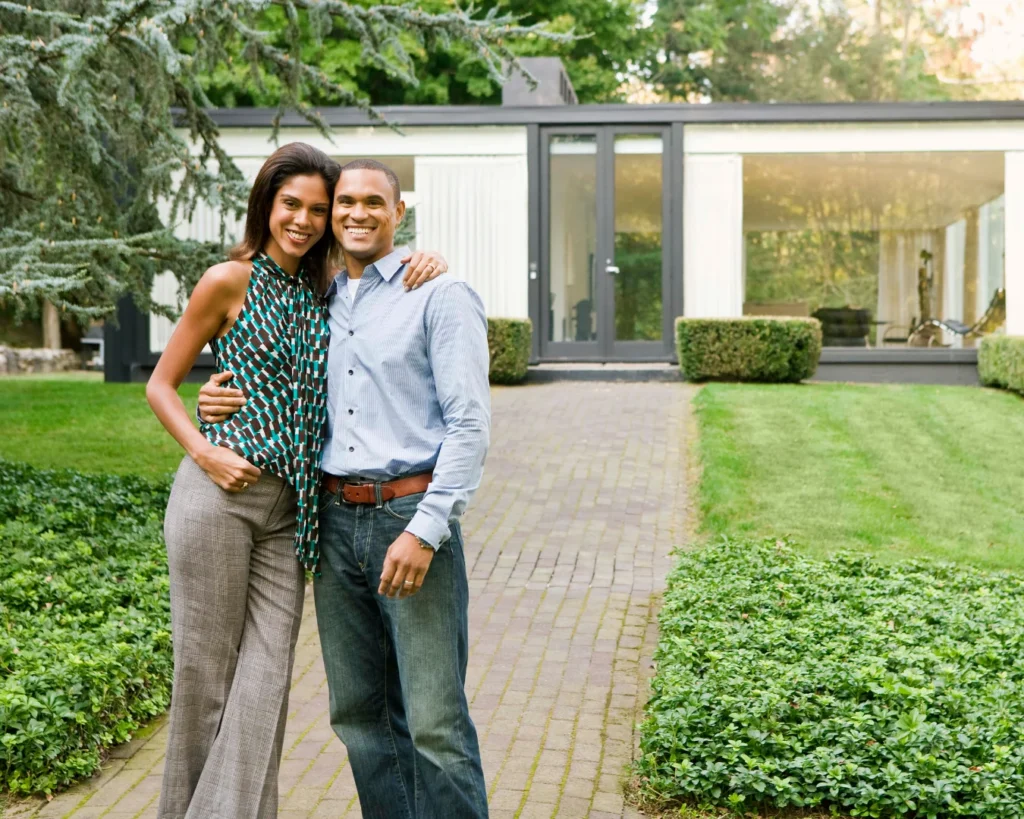 A smiling couple stands arm in arm on a paved path outside a modern home, with trimmed green hedges and a lawn nearby.