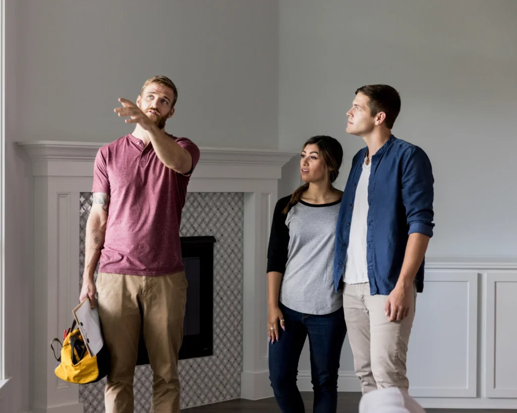 A bearded man in a pink shirt gestures upward while a woman and another man look toward the ceiling in a living room.