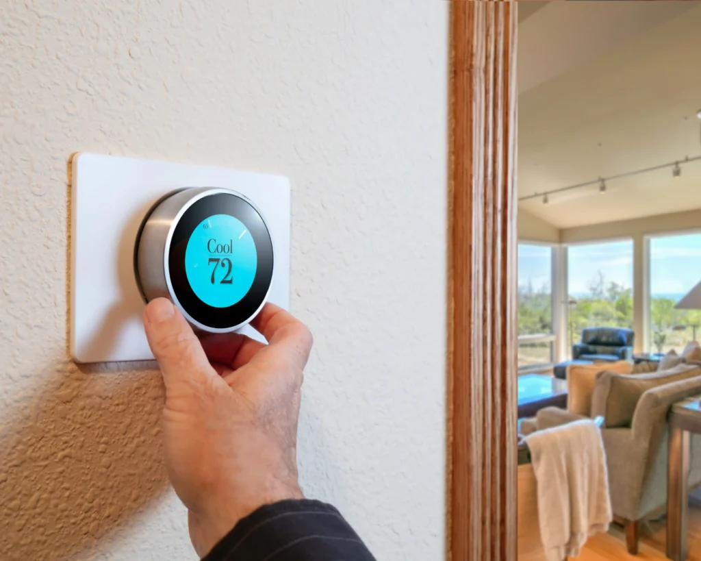 A hand adjusts a digital thermostat set to 72 degrees beside a bright living room with large windows and comfortable seating.