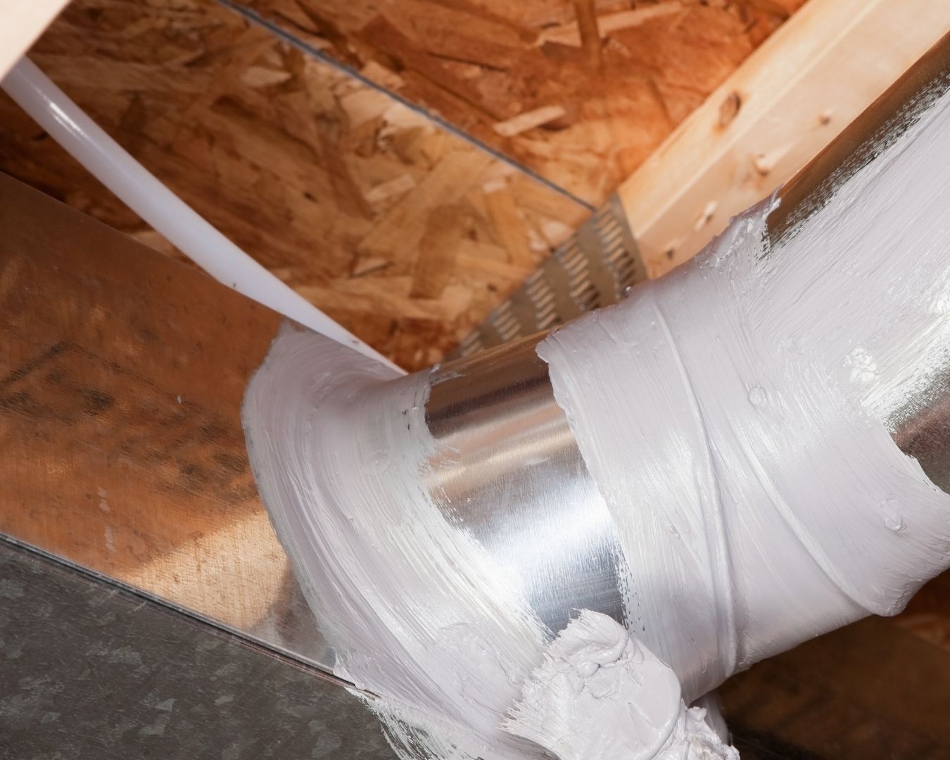 White HVAC ducts meet at a broad wrapped joint beneath exposed brown roof sheathing in an unfinished attic.