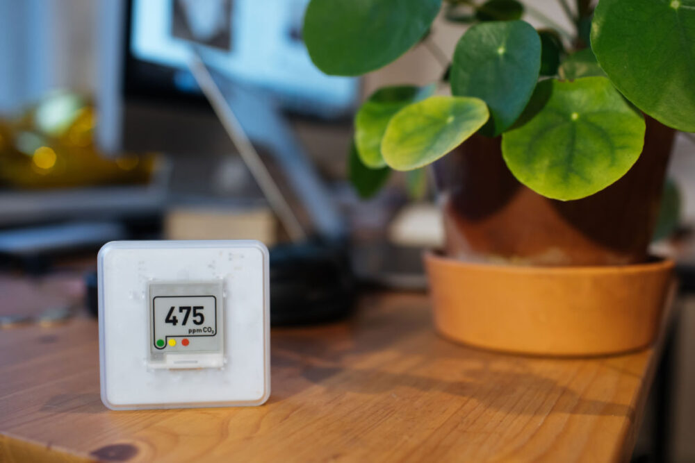 An air quality measuring device rests on a wooden table next to a green-leafed plant in a pot.