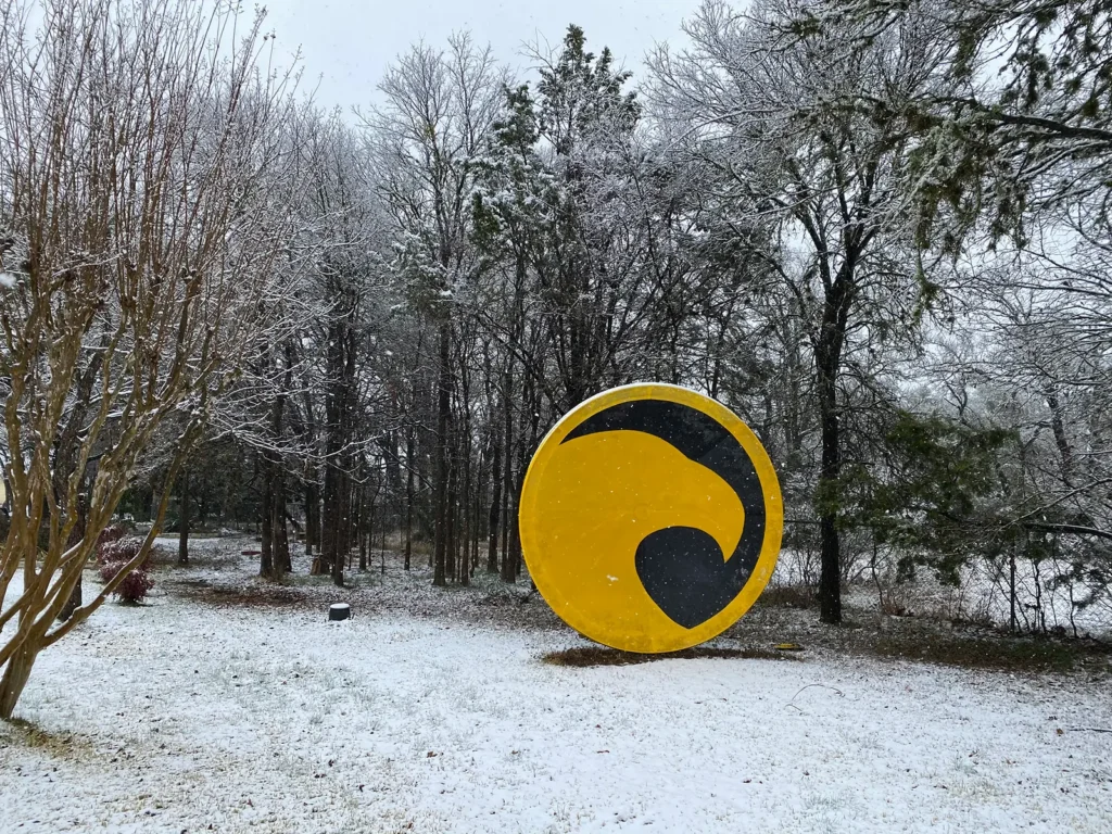 A golden circular eagle emblem stands in a snow-covered yard, framed by leafless trees and dark evergreens beneath a pale winter sky.