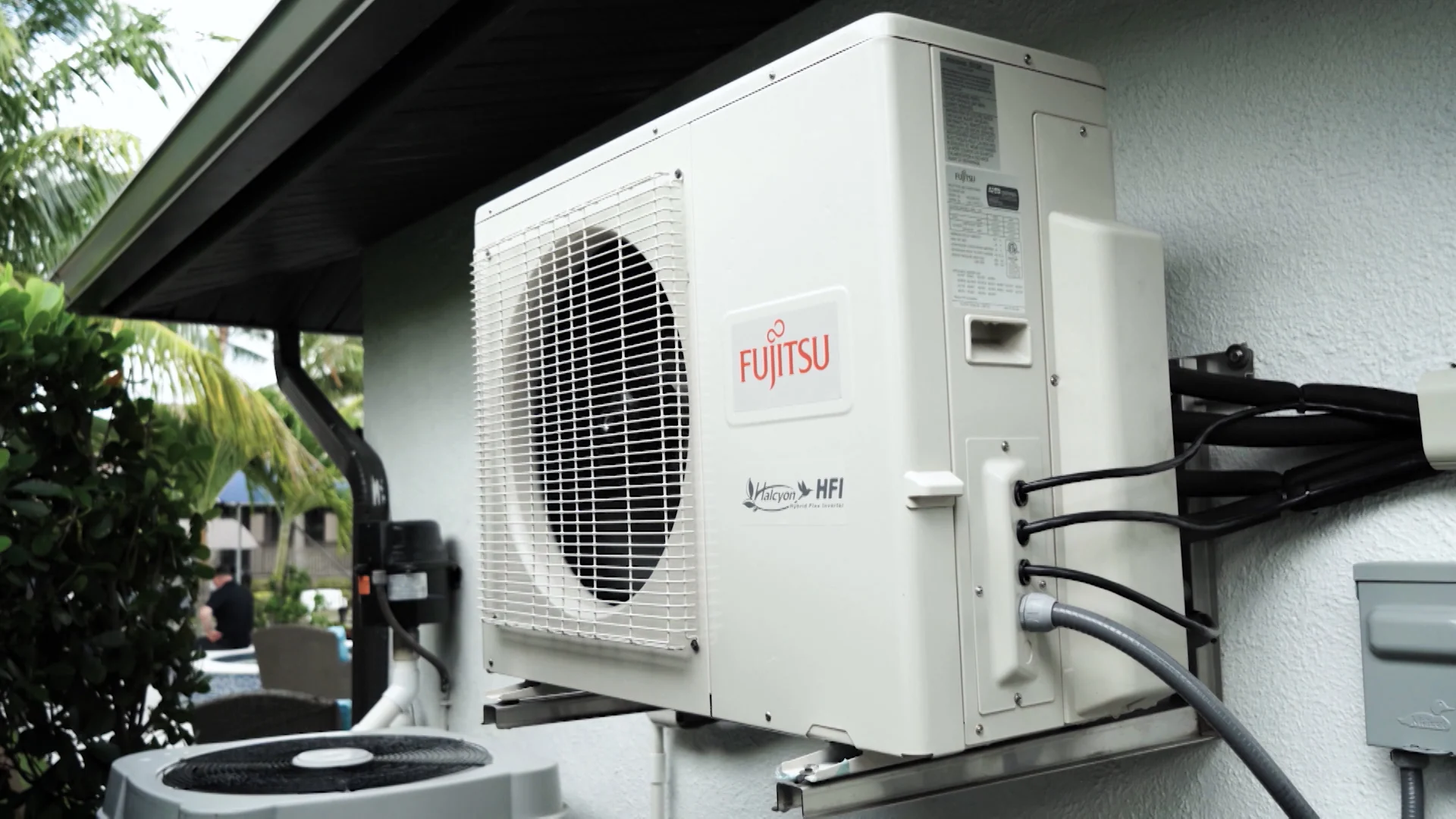 A white Fujitsu ductless HVAC unit is mounted on an exterior wall, with a broad grille, dark fan, and connected lines visible.