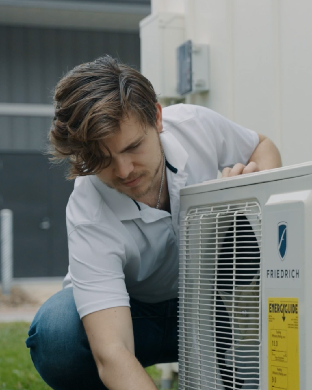 A technician in a white collared shirt leans over a Frigidaire air conditioning unit, inspecting its front grille and energy label.
