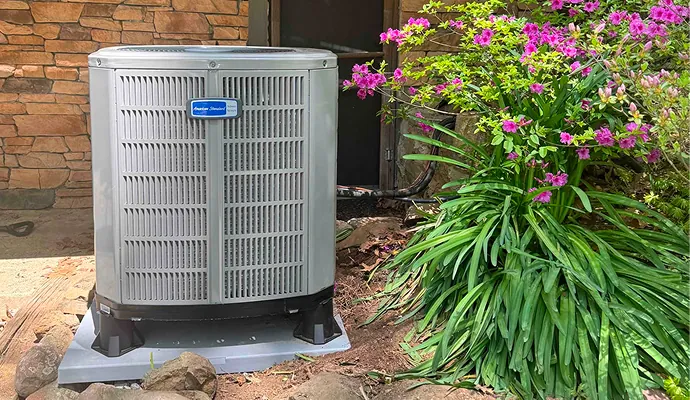 A gray American Standard outdoor air conditioner stands beside a stone wall, framed by green foliage and bright pink flowers.