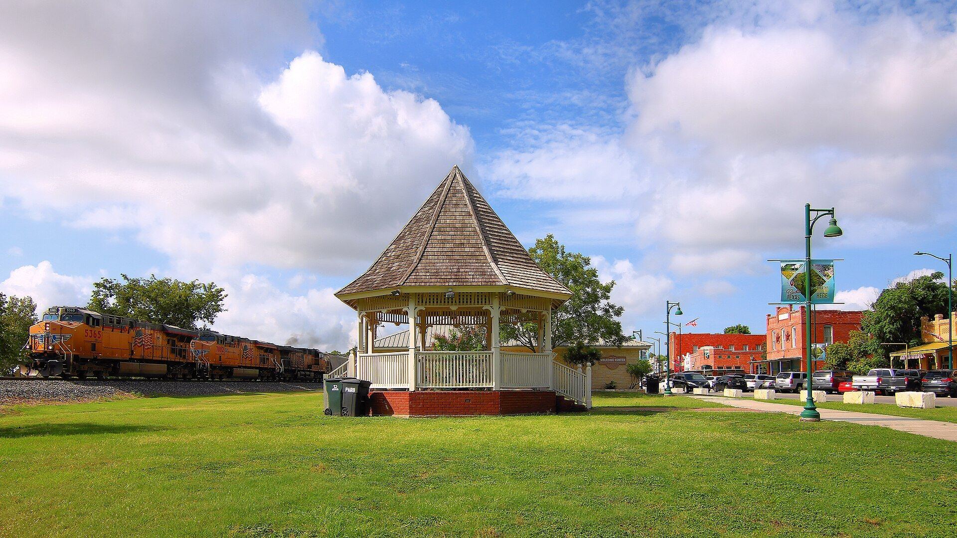 White gazebo on the green in historic downtown Buda, Texas, with a freight train and Main Street buildings behind