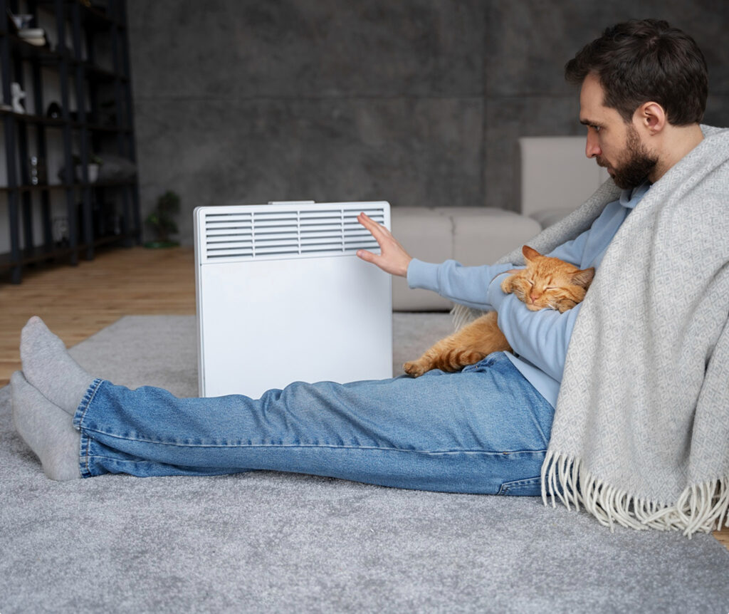 A seated man in a blue shirt reaches toward a portable air purifier while an orange cat rests on his lap in a living room.