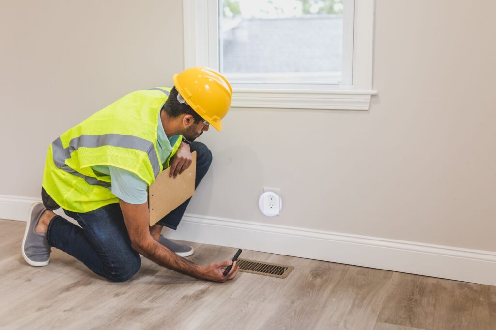 Contractor in a hard hat inspecting a floor air vent in a home
