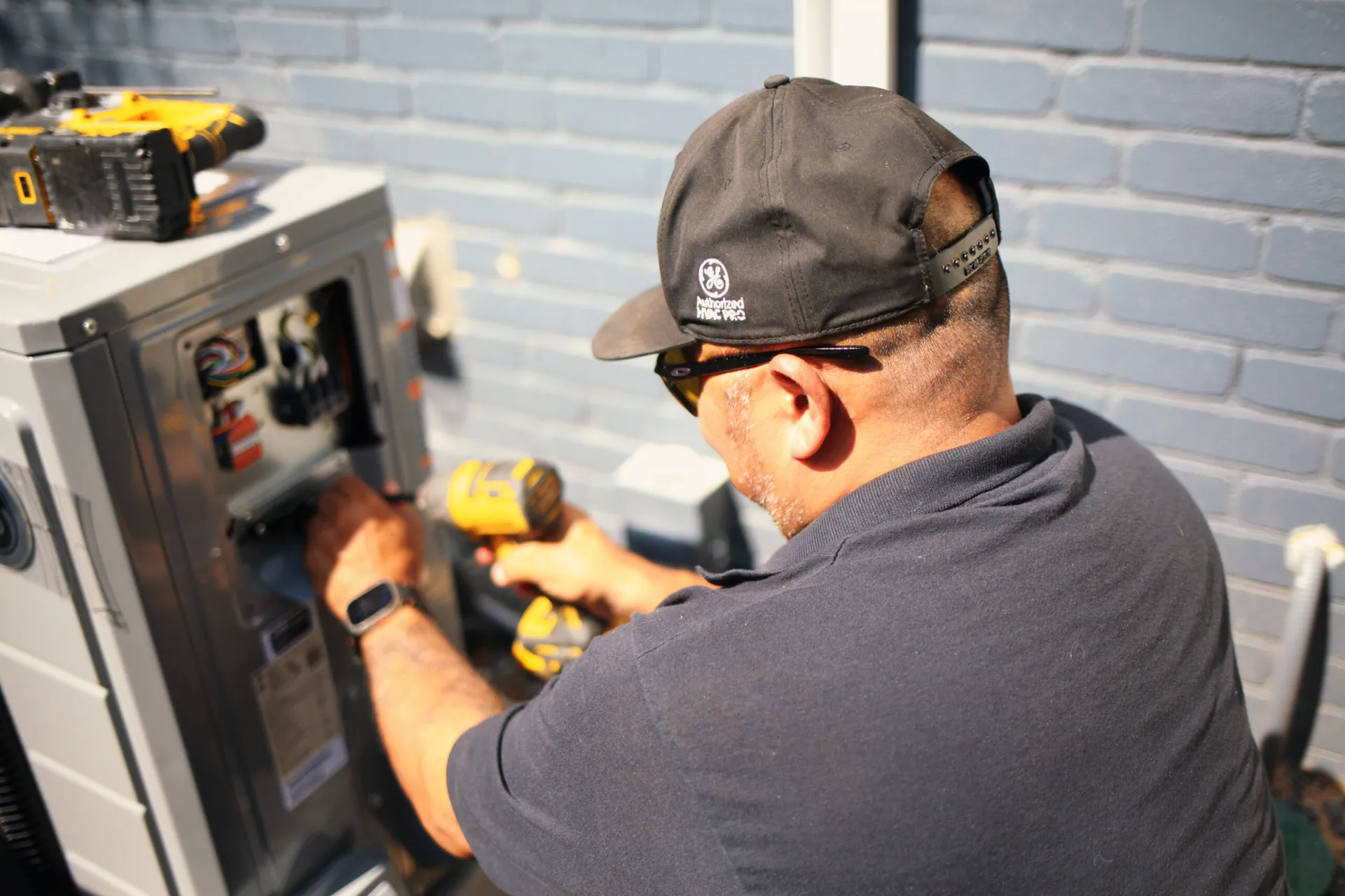 A technician in a dark cap and safety glasses uses a yellow cordless drill while working inside an outdoor HVAC unit's open panel.