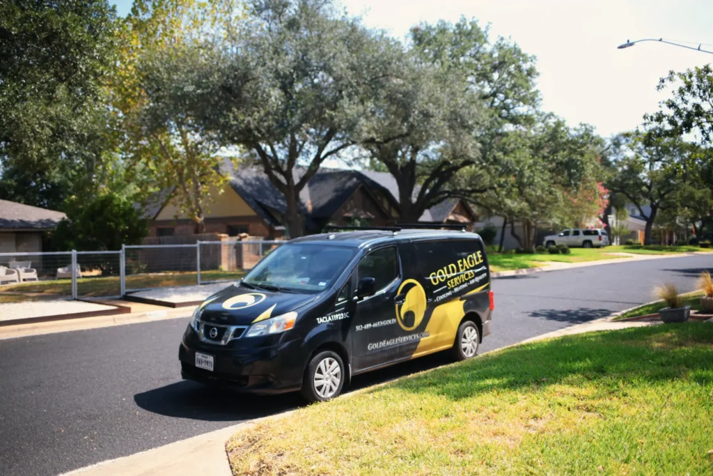 A black and gold Gold Eagle Services HVAC van is parked beside a sunny residential street lined with trees and homes.