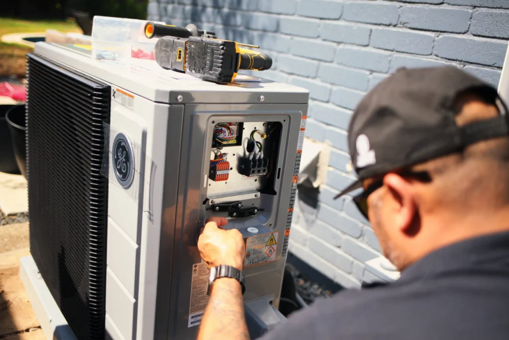 A technician adjusts exposed wiring inside a gray outdoor heat pump, with a cordless drill on top beside a light blue brick wall.