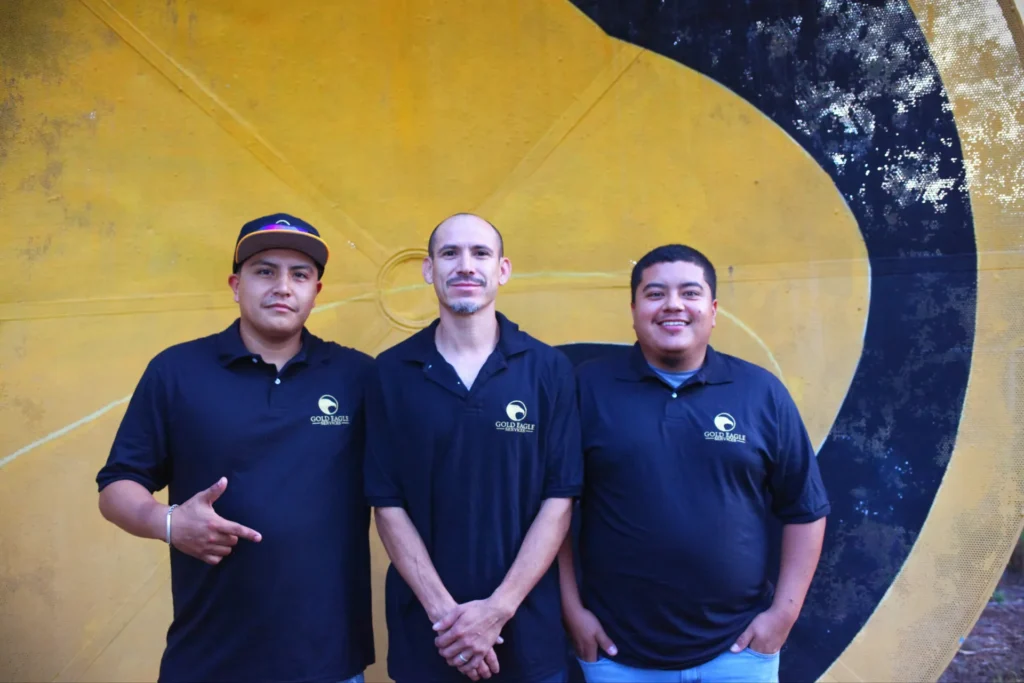 Three Gold Eagle Services employees stand together in navy polo shirts before a large yellow and black circular backdrop.