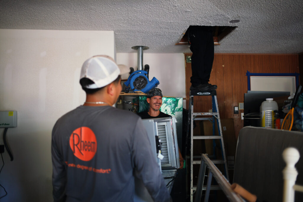 A heating technician in a gray shirt and white cap works beside HVAC equipment as a coworker stands near an open ceiling access.