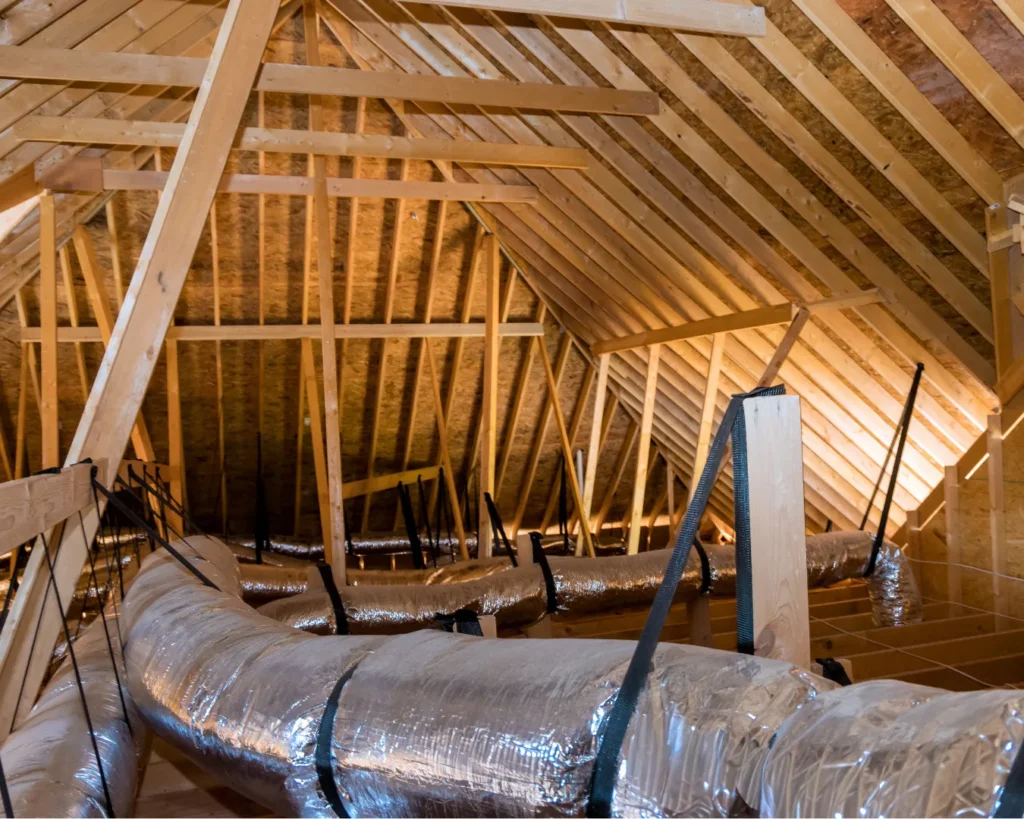 Silver insulated HVAC ducts run beneath exposed wooden attic rafters, secured by dark straps across the ventilation system.