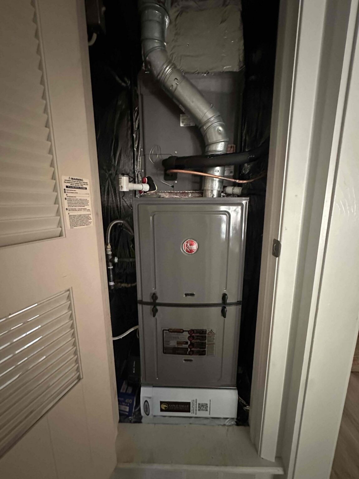 Upflow gas furnace in a closet with a louvered door, a metal vent pipe and a coil cabinet above