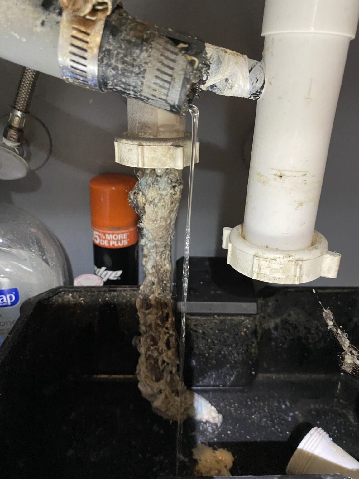 Under-sink view of a condensate line ending at a utility sink, with a rope of slime hanging from the fitting into the basin