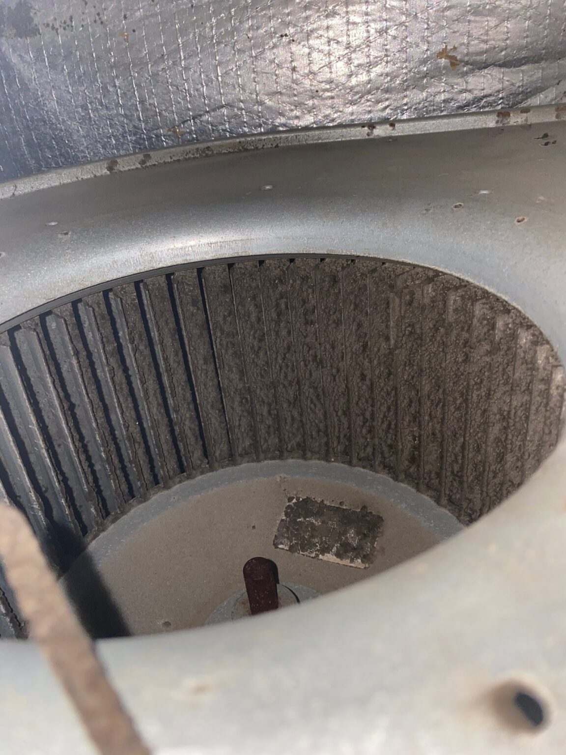 Looking down into a blower wheel: its blades are caked with gray dirt, with a clump of debris on the wheel's center plate beside the motor shaft