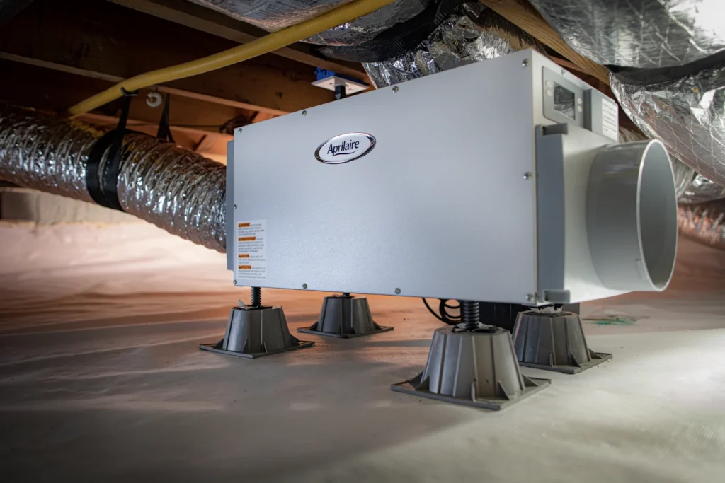 A gray Aprilaire whole-home dehumidifier rests on supports among insulated HVAC ducts in a residential attic.