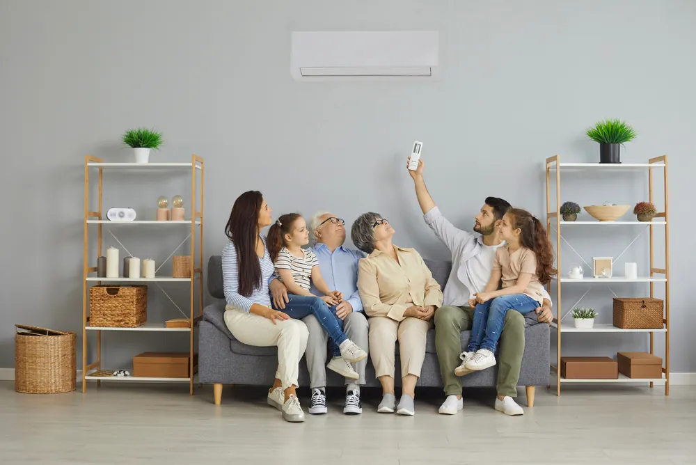 A family of three generations (mother and father, two little girls, and grandparents) sitting on a gray couch as the dad uses a remote to turn on the zoned HVAC system