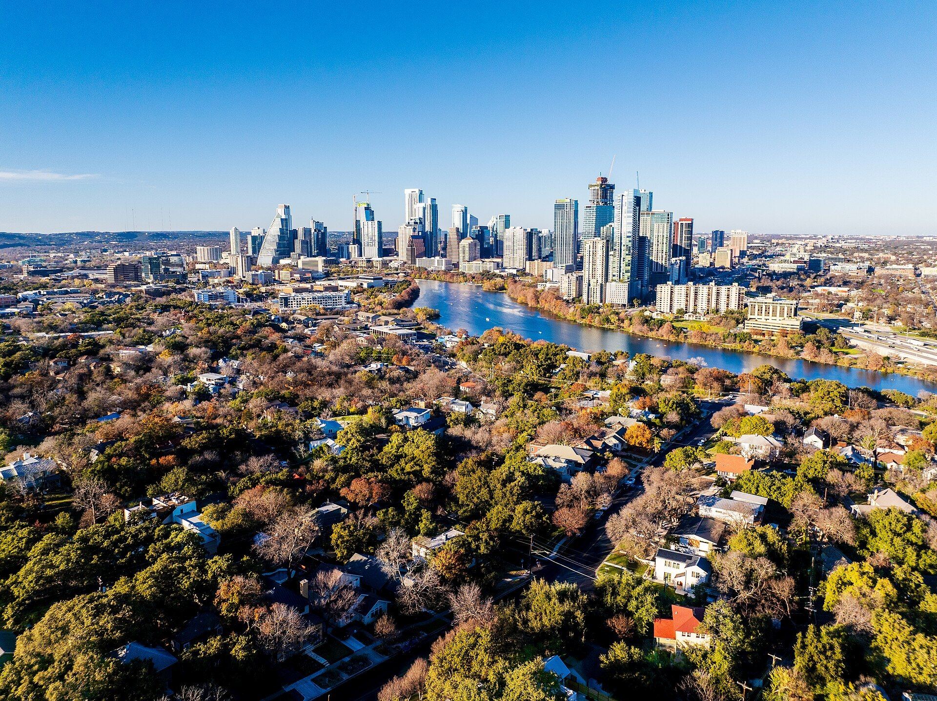 Aerial view over the tree-covered Travis Heights neighborhood toward Lady Bird Lake and the downtown Austin skyline