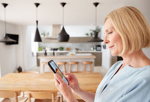 A smiling woman holds a cellphone that controls a smart thermostat while standing in her modern kitchen.