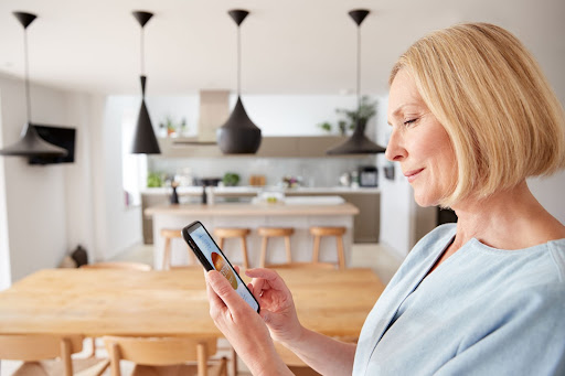 A smiling woman holds a cellphone that controls a smart thermostat while standing in her modern kitchen.