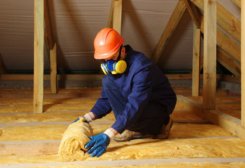 A person wearing blue gloves, a blue jacket, a colorful gas mask, and an orange hard hat rolls up insulation in an attic space.