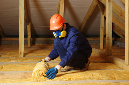 A person wearing blue gloves, a blue jacket, a colorful gas mask, and an orange hard hat rolls up insulation in an attic space.